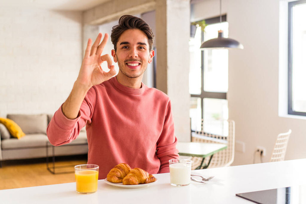 Young mixed race man having breakfast in a kitchen on the morning cheerful and confident showing ok gesture. - Photo, Image