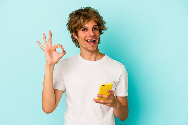 Jonge blanke man met make-up houden mobiele telefoon geïsoleerd op blauwe achtergrond vrolijk en vol vertrouwen tonen ok gebaar. - Foto, afbeelding