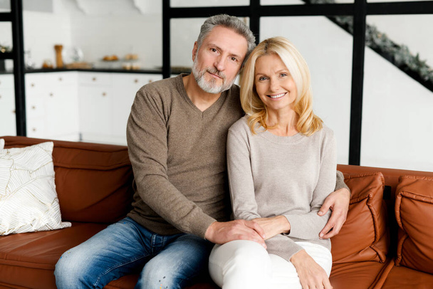 Middle-aged couple in love. Cheerful elderly couple smiling and hugging at cozy living room, spends time together in cozy living room. Portrait of senior spouses looking at the camera and smile - Zdjęcie, obraz
