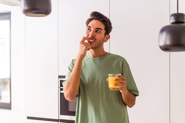 Jonge gemengde ras man drinken sinaasappelsap in zijn keuken ontspannen denken over iets op zoek naar een kopieerruimte. - Foto, afbeelding