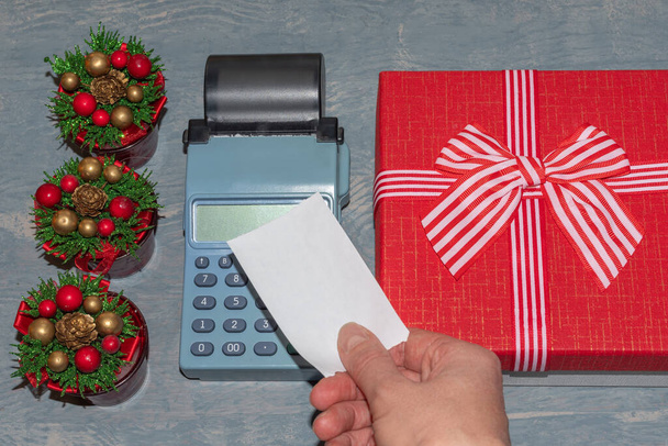 Christmas sale. A hand holding a Blank white check above the cash register near a red gift box with a ribbon and little Christmas trees with cones. Buying gifts for New Year and Christmas - Фото, зображення