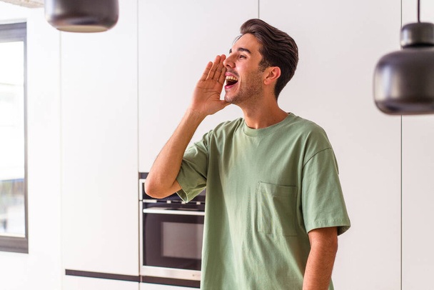 Young mixed race man in his kitchen shouting and holding palm near opened mouth. - Photo, image
