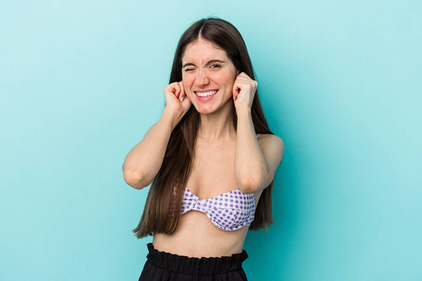 Young caucasian woman wearing bikini isolated on blue background covering ears with hands. - 写真・画像