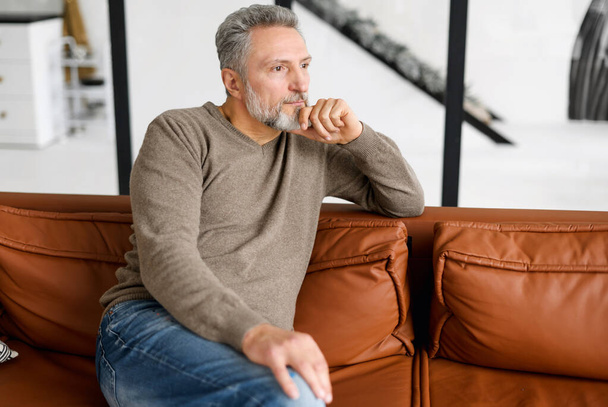 Portrait of thoughtful mid adult man sitting on the leather sofa at home - Foto, Imagen