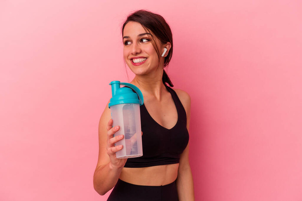 Young caucasian woman drinking a protein shake isolated on pink background looks aside smiling, cheerful and pleasant. - Photo, Image