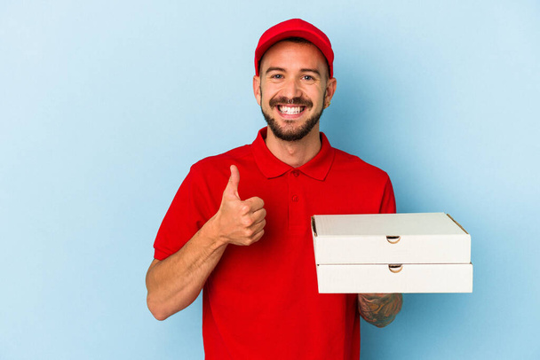 Junger kaukasischer Lieferant mit Tätowierungen, der Pizzen isoliert auf blauem Hintergrund hält, lächelt und den Daumen nach oben hebt - Foto, Bild