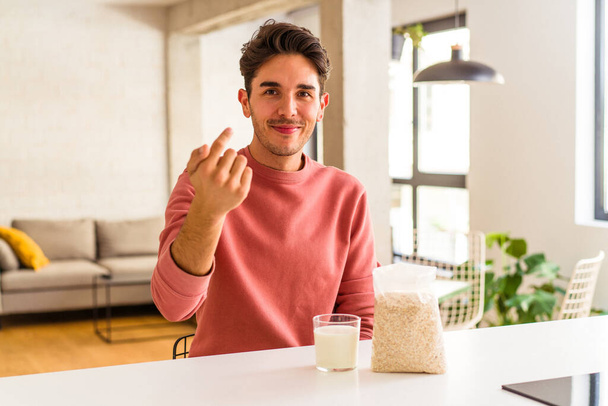 Νεαρός άνδρας μικτής φυλής τρώει πλιγούρι βρώμης και γάλα για πρωινό στην κουζίνα του δείχνοντάς σας με το δάχτυλο σαν να σας προσκαλεί να έρθετε πιο κοντά. - Φωτογραφία, εικόνα