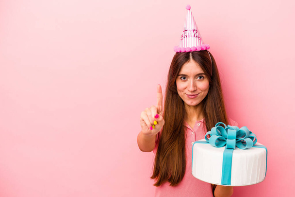 Jeune femme caucasienne célébrant un anniversaire isolé sur fond rose montrant numéro un avec doigt. - Photo, image