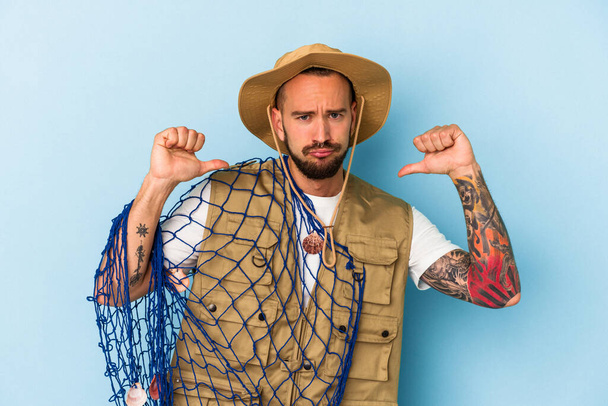 Joven pescador caucásico con tatuajes sosteniendo red aislada sobre fondo azul se siente orgulloso y seguro de sí mismo, ejemplo a seguir. - Foto, Imagen