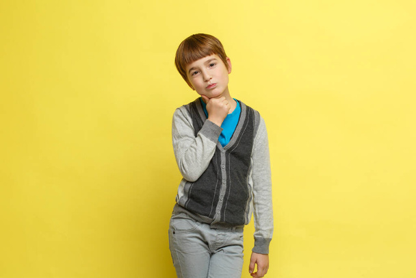 Cute doubting boy in casual stylish clothes holds his hand at the chin stands isolated on a yellow background. - Fotoğraf, Görsel