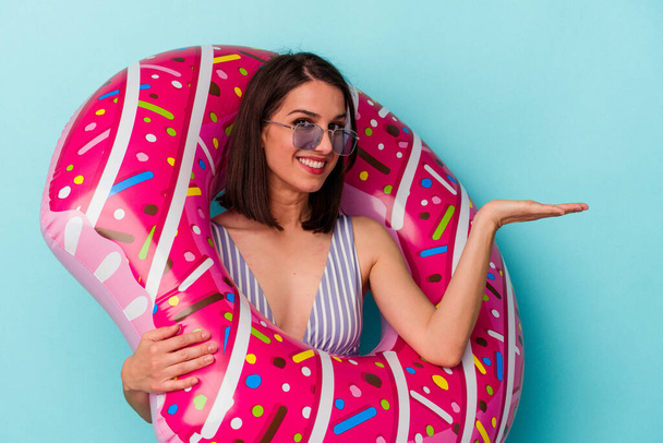 Young caucasian woman with inflatable donut isolated on blue background showing a copy space on a palm and holding another hand on waist. - Photo, Image