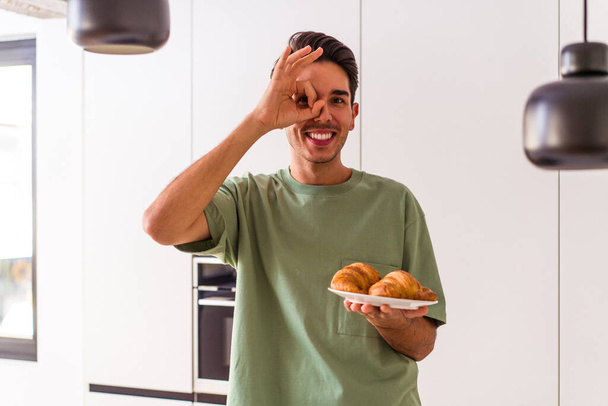 Νέοι μικτή φυλή άνθρωπος τρώει κρουασάν σε μια κουζίνα το πρωί ενθουσιασμένος κρατώντας εντάξει χειρονομία στο μάτι. - Φωτογραφία, εικόνα