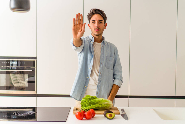 Jeune homme métis préparant une salade pour le déjeuner debout avec la main tendue montrant le signe d'arrêt, vous empêchant. - Photo, image