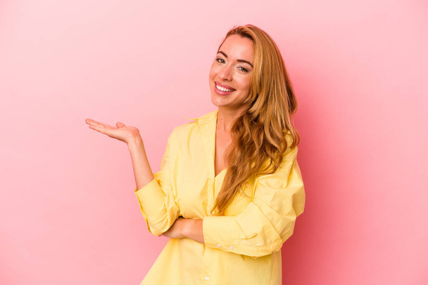 Caucasian blonde woman isolated on pink background showing a copy space on a palm and holding another hand on waist. - Foto, imagen