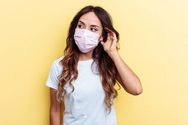 Young mixed race woman wearing a protection for virus isolated on yellow background Young mixed race woman wearing a protection for virus isolated on yellow background trying to listening a gossip. - Фото, изображение