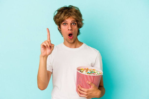 Jonge blanke man met make-up houden popcorn geïsoleerd op blauwe achtergrond met een idee, inspiratie concept. - Foto, afbeelding