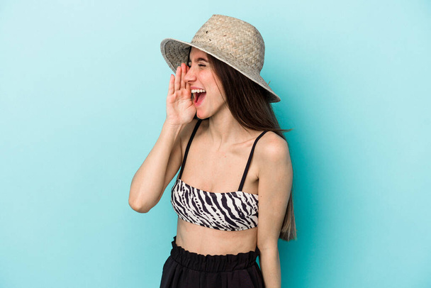 Jonge blanke vrouw die naar het strand gaat en bikini draagt geïsoleerd op een blauwe achtergrond schreeuwend en handpalm vasthoudend bij geopende mond. - Foto, afbeelding