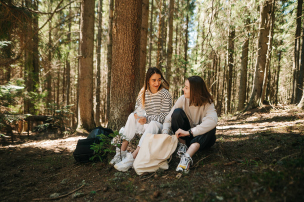 Női barátok pihennek az erdőben a szabadban. Két gyönyörű nő turista üldögél a hegyi erdőben és pihen, mosollyal az arcukon beszélgetve.. - Fotó, kép