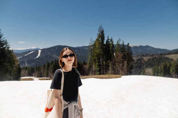 Beautiful young woman hiker in stylish casual clothes and a shoulder bag posing for the camera on a snowy mountain in sunny weather on the background of an incredible landscape. - Фото, изображение