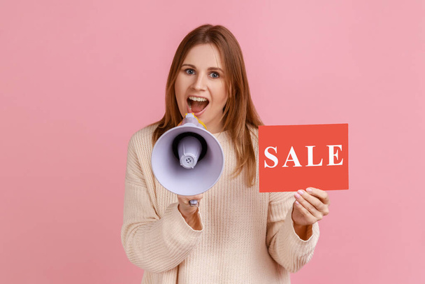 Portrait of excited young adult blond woman holding card with sale inscription and screaming in megaphone about discounts, wearing white sweater. Indoor studio shot isolated on pink background. - Foto, immagini