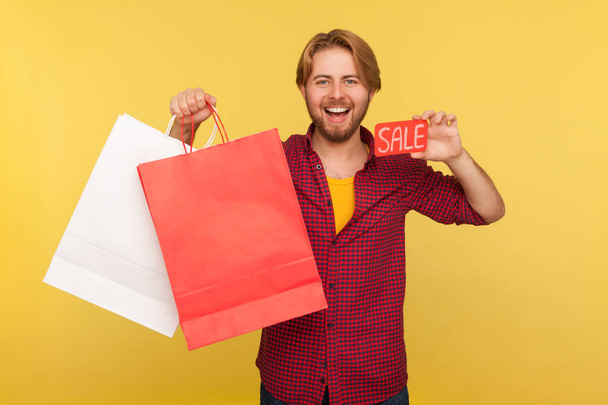 Portrait of hipster bearded guy in checkered shirt looking at camera with excitement, holding shopping bags and card with sale inscription. Indoor studio shot isolated on yellow background. - Фото, изображение