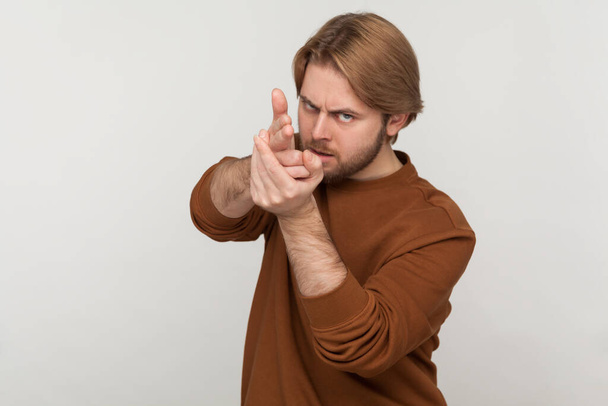 Portrait of dangerous young adult handsome man with beard wearing sweatshirt, pointing with finger pistols at camera, pretending to shoot. Indoor studio shot isolated on gray background. - Fotografie, Obrázek