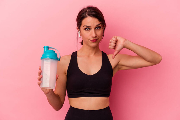 Young caucasian woman drinking a protein shake isolated on pink background showing a dislike gesture, thumbs down. Disagreement concept. - Photo, Image