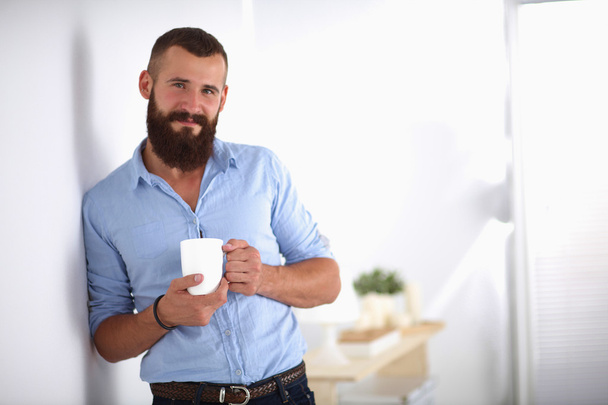 Young man  standing near wall and holding cup of coffee in offi - Фото, изображение