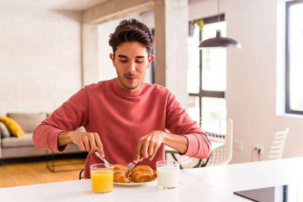 Young mixed race man eating croissant in a kitchen on the morning - 写真・画像