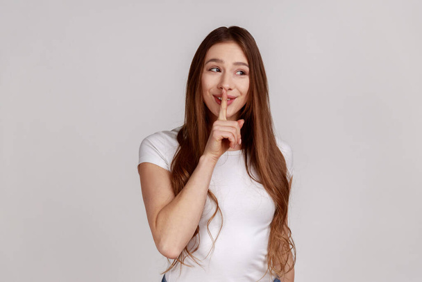 Shh, it's big secret. Beautiful cunning woman smiling, showing gesture secret sign with finger near her lips, wearing white T-shirt. Indoor studio shot isolated on gray background. - Fotó, kép