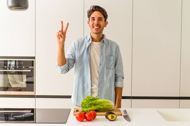 Jonge gemengde ras man bereidt een salade voor lunch tonen nummer twee met vingers. - Foto, afbeelding