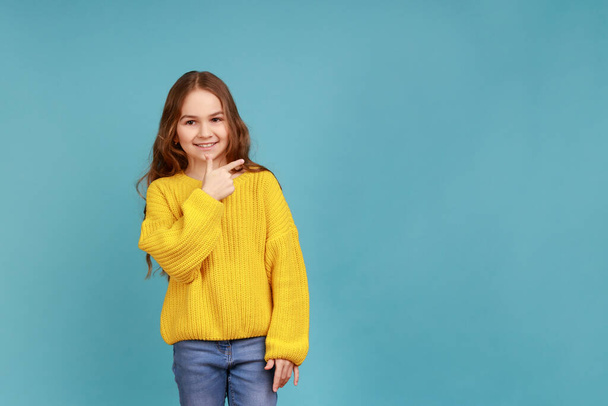 Little girl pointing to empty place on background, preschooler showing copy space for promotion, wearing yellow casual style sweater. Indoor studio shot isolated on blue background. - Photo, Image
