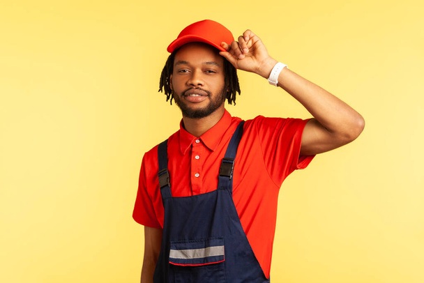 Portrait of attractive bearded handyman wearing uniform standing with raised arm, keeps hand on his visor cap, waiting orders in service industry. Indoor studio shot isolated on yellow background. - Fotó, kép