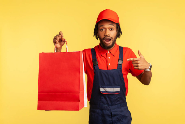 Astonished shocked deliveryman wearing blue overalls and red T-shirt pointing finger at shopping bags, very fast delivery service. Indoor studio shot isolated on yellow background. - Fotografie, Obrázek