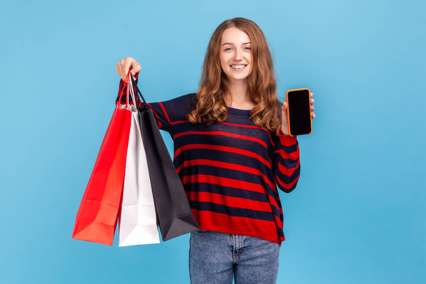 Happy woman wearing striped casual style sweater, standing with satisfied look showing shopping bags and smart phone with blank screen for ad. Indoor studio shot isolated on blue background. - Foto, immagini