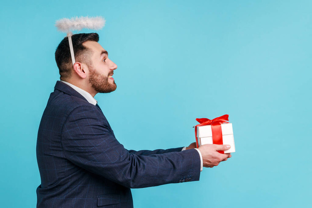 Side view of handsome bearded male wearing official style suit, halo over head giving gift box, offering present on holiday, looking ahead. Indoor studio shot isolated on blue background. - 写真・画像