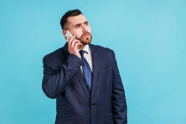 Serious bearded man wearing dark official style suit holding phone near his ear, talks to his companies, solving important work issues. Indoor studio shot isolated on blue background. - Fotoğraf, Görsel