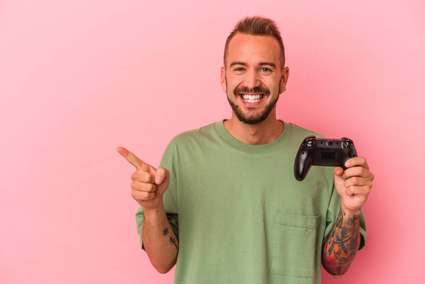 Jovem caucasiano com tatuagens segurando controlador de jogo isolado em fundo rosa sorrindo e apontando para o lado, mostrando algo no espaço em branco. - Foto, Imagem