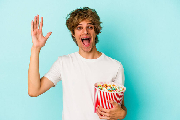 Fiatal fehér férfi smink gazdaság popcorn elszigetelt kék háttérrel kap egy kellemes meglepetés, izgatott és felemeli a kezét. - Fotó, kép