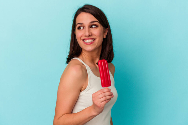 Young caucasian woman holding an ice cream isolated on blue background looks aside smiling, cheerful and pleasant. - Foto, imagen