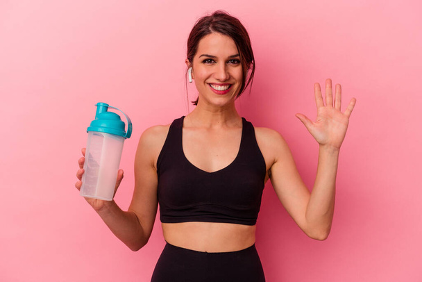Joven mujer caucásica bebiendo un batido de proteínas aislado sobre fondo rosa sonriente alegre mostrando el número cinco con los dedos. - Foto, Imagen