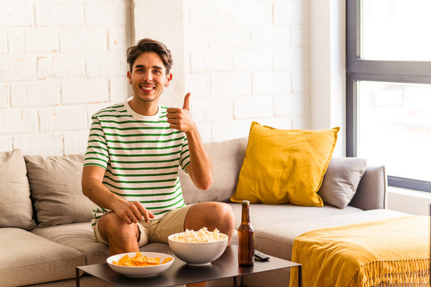 Молодой человек смешанной расы ест попкорны сидя на диване улыбаясь и поднимая большой палец вверх - Фото, изображение