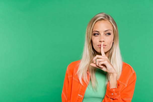 knifflige blonde Frau zeigt Schweigezeichen, während sie isoliert auf Grün zur Seite schaut - Foto, Bild