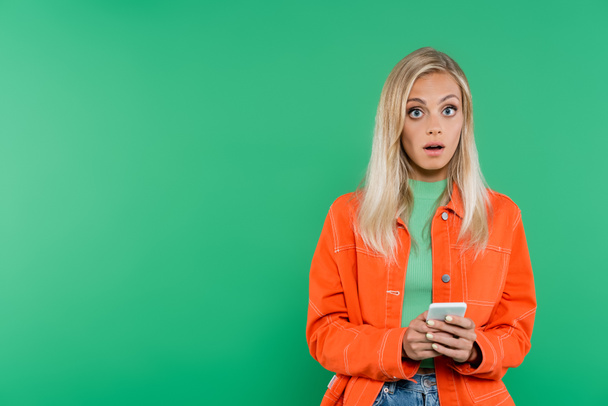 překvapená blondýna žena v oranžové bundě při pohledu na kameru, zatímco chatování na mobil izolované na zelené - Fotografie, Obrázek
