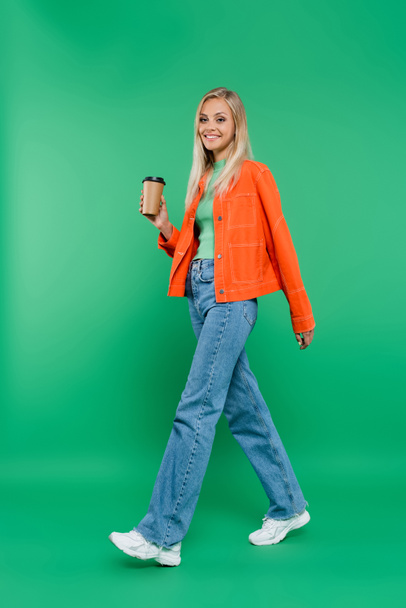 full length view of happy young woman in jeans and orange jacket walking with takeaway drink on green - Photo, Image