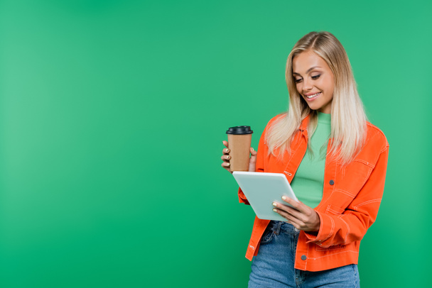 gelukkig blond vrouw met afhaaldrankje kijken naar digitale tablet geïsoleerd op groen - Foto, afbeelding