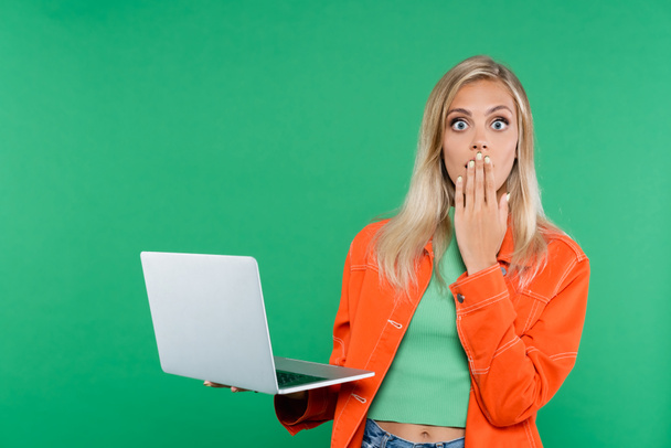 nervous blonde woman covering mouth with hand while holding laptop isolated on green - Foto, Bild