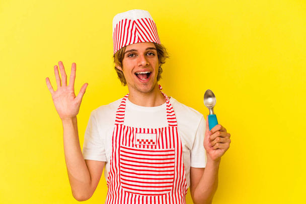 Nuori jäätelö maker mies meikki tilalla lusikka eristetty keltaisella taustalla hymyilevä iloinen osoittaa numero viisi sormin. - Valokuva, kuva
