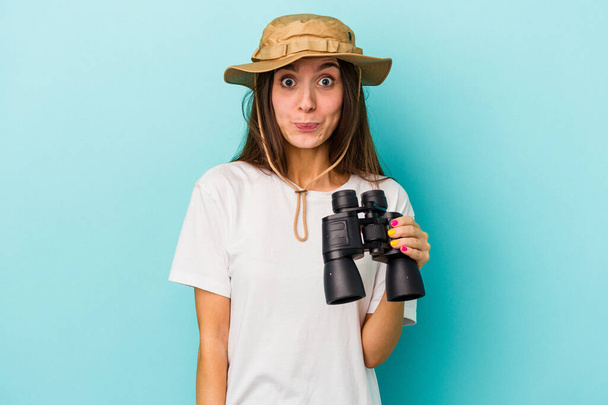 Young caucasian explorer woman holding binoculars isolated on blue background shrugs shoulders and open eyes confused. - Foto, afbeelding