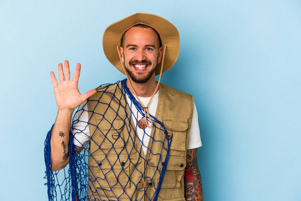 Joven pescador caucásico con tatuajes sosteniendo red aislada sobre fondo azul sonriente alegre mostrando el número cinco con los dedos. - Foto, Imagen